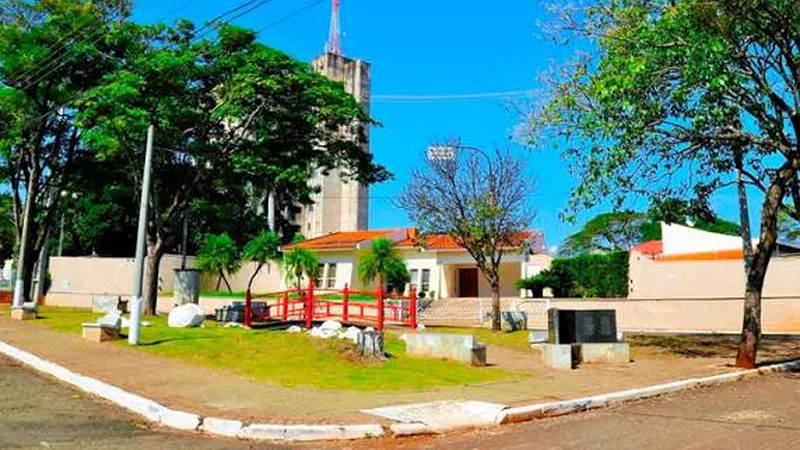 Praça arborizada com ponte vermelha e torre ao fundo em Sertaneja, Paraná